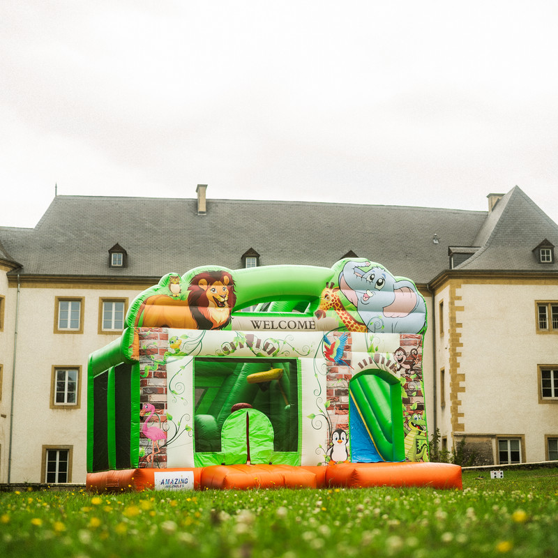 Grand château gonflable Zoo vert avec lion, éléphant et toboggan devant un château luxembourgeois.